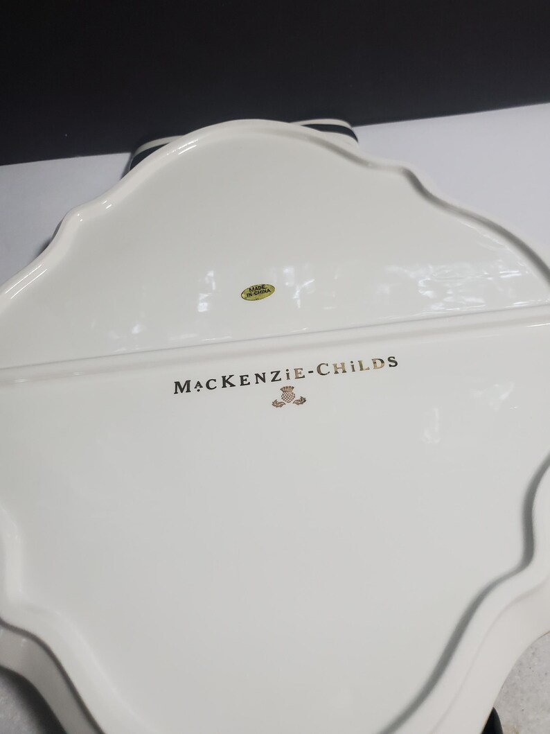 May include: The underside of a white ceramic serving tray with a scalloped edge. The text MACKENZIE-CHILDS is printed in black lettering above a small gold emblem. A circular yellow sticker with the text MADE IN CHINA is visible near the top.