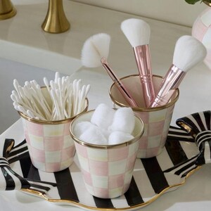 May include: Three cylindrical containers with a pink and white checkered pattern and gold rims, holding cotton swabs, cotton balls, and makeup brushes with rose gold handles. These sit on a black and white striped tray with gold-trimmed edges.