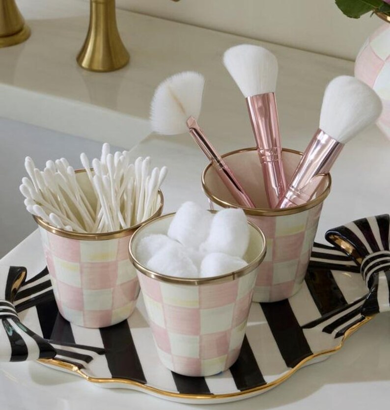 May include: Three cylindrical containers with a pink and white checkered pattern and gold rims, holding cotton swabs, cotton balls, and makeup brushes with rose gold handles. These sit on a black and white striped tray with gold-trimmed edges.
