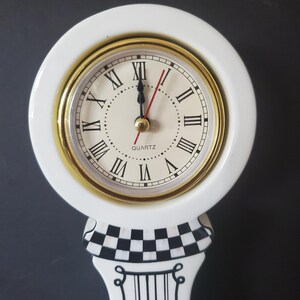 May include: A white quartz clock with a gold-tone bezel and Roman numeral hour markers. The clock sits atop a decorative stand with a black and white checkered pattern and vertical black stripes.