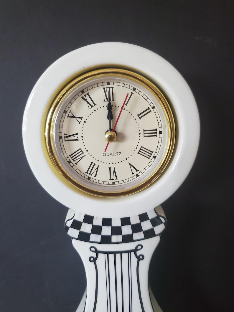 May include: A white quartz clock with a gold-tone bezel and Roman numeral hour markers. The clock sits atop a decorative stand with a black and white checkered pattern and vertical black stripes.
