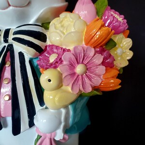 Puede incluir: Una figura de conejo blanco con una pajarita a rayas blancas y negras, camisa rosa y un ramo de flores coloridas. El ramo incluye flores rosas, naranjas, amarillas y moradas, y un peque&ntilde;o pollito amarillo. La figura tiene una expresi&oacute;n alegre.