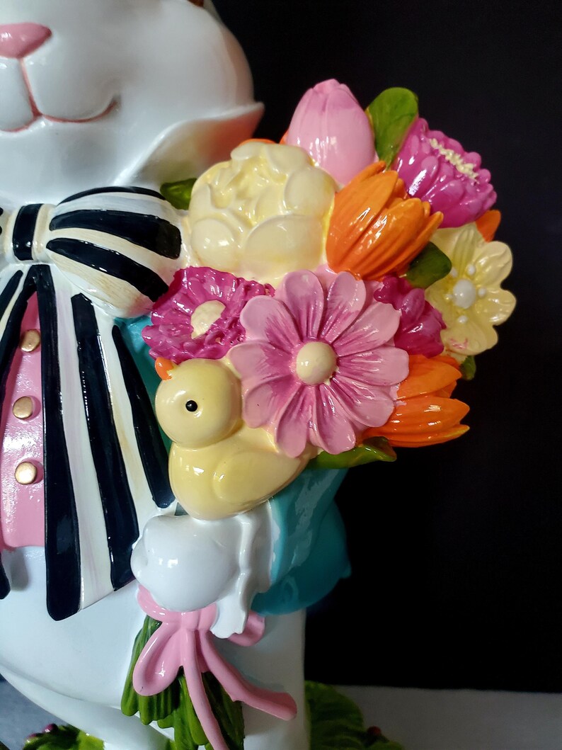 Puede incluir: Una figura de conejo blanco con una pajarita a rayas blancas y negras, camisa rosa y un ramo de flores coloridas. El ramo incluye flores rosas, naranjas, amarillas y moradas, y un peque&ntilde;o pollito amarillo. La figura tiene una expresi&oacute;n alegre.