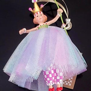 May include: A whimsical figurine of a princess in a layered tulle dress with pink, purple, and blue hues. She wears a gold crown and holds a wand with a pink and blue top. Her checkered pants and pink shoes complete the look.