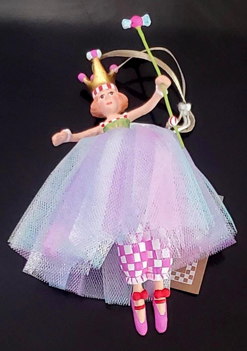 May include: A whimsical figurine of a princess in a layered tulle dress with pink, purple, and blue hues. She wears a gold crown and holds a wand with a pink and blue top. Her checkered pants and pink shoes complete the look.