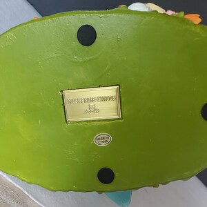 Puede incluir: La parte inferior de una base de figura decorativa verde con cuatro pies circulares negros. Una placa rectangular dorada dice "Mackenzie Childs" con un logotipo de oso de peluche. Una peque&ntilde;a etiqueta dice "Made in China."
