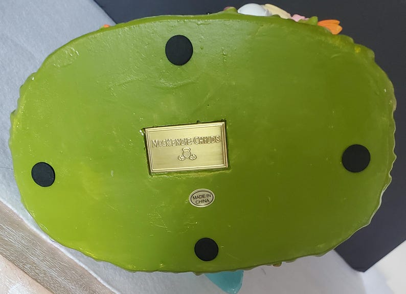 Puede incluir: La parte inferior de una base de figura decorativa verde con cuatro pies circulares negros. Una placa rectangular dorada dice "Mackenzie Childs" con un logotipo de oso de peluche. Una peque&ntilde;a etiqueta dice "Made in China."