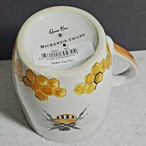 May include: A white ceramic mug featuring a painted bee with a black and white checkered pattern on its body, surrounded by yellow honeycomb illustrations. The base displays the text Queen Bee, MACKENZIE-CHILDS, and a barcode sticker.