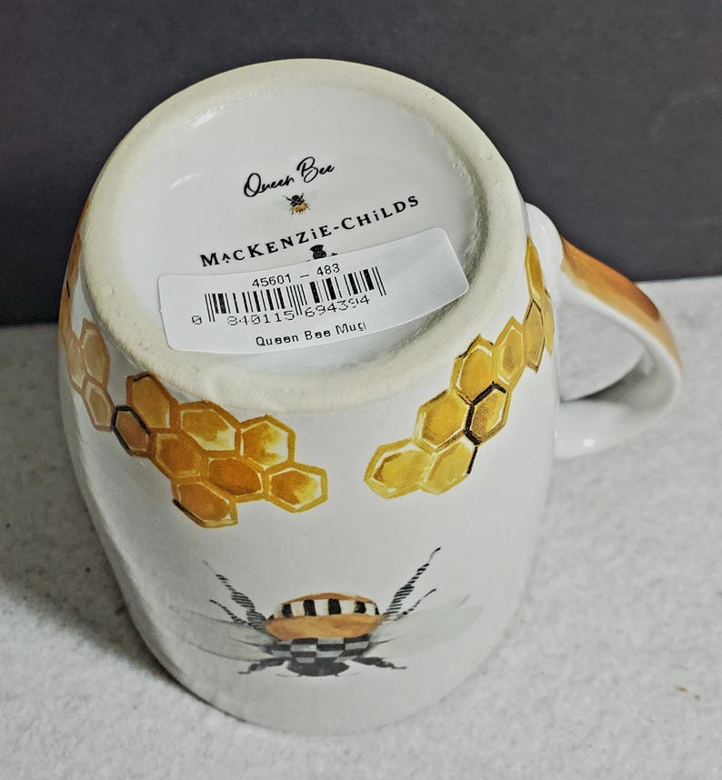 May include: A white ceramic mug featuring a painted bee with a black and white checkered pattern on its body, surrounded by yellow honeycomb illustrations. The base displays the text Queen Bee, MACKENZIE-CHILDS, and a barcode sticker.