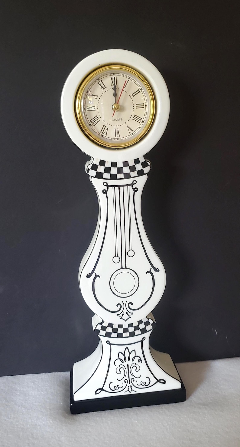 May include: A white quartz clock with a gold-tone bezel and Roman numerals. The clock has a decorative black and white checkered and scrollwork design. The clock is approximately 12 inches tall.