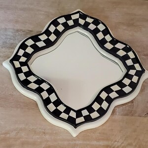 Puede incluir: Un espejo de pared con forma de cuatrifolio que presenta un borde con patr&oacute;n de cuadros en blanco y negro. El marco tiene un acabado liso y brillante con un borde exterior blanco y una superficie de vidrio reflectante en el centro.