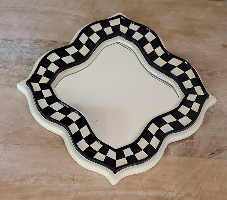 Puede incluir: Un espejo de pared con forma de cuatrifolio que presenta un borde con patr&oacute;n de cuadros en blanco y negro. El marco tiene un acabado liso y brillante con un borde exterior blanco y una superficie de vidrio reflectante en el centro.