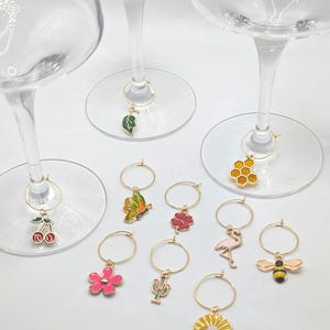 Peut inclure: Ensemble de breloques de vin dorées avec divers motifs colorés, dont une cerise, un papillon, une fleur, un cactus, un flamant rose, une abeille et un nid d'abeilles. Les breloques sont suspendues aux pieds de verres à vin clairs.