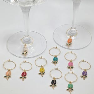 Peut inclure: Un assortiment de charmants breloques à vin en forme de tulipe. Chaque breloque est composée d'un anneau doré orné d'un pendentif tulipe de différentes couleurs, comme le rose, le jaune et le violet. Les breloques sont présentées près de verres à vin.