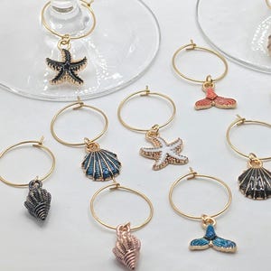 Peut inclure: Un assortiment de breloques de verre à vin dorées avec des pendentifs sur le thème de l'océan. Les breloques comprennent des étoiles de mer, des coquillages et une queue de sirène de différentes couleurs comme le noir, le bleu et le rose. Les breloques sont conçues pour être accrochées à la tige d'un verre à vin.