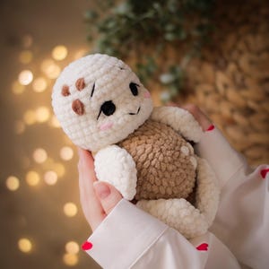 May include: A plush crochet turtle toy featuring a cream-colored head with brown spots, black embroidered eyes, and pink cheeks. The turtle has a textured, light brown rounded shell and cream-colored limbs made from soft, fuzzy yarn material.