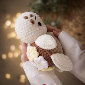 May include: A plush crochet turtle toy featuring a cream-coloured head with brown embroidered details, a brown textured shell, and cream-coloured flippers. The shell is decorated with small, soft, white and pale yellow crocheted flower appliques.