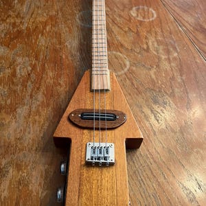 Könnte beinhalten: Eine handgefertigte Zigarrenkistengitarre aus Holz mit dreieckigem Korpus und langem Hals. Das Instrument verfügt über ein Griffbrett aus Holz, Metall-Stimmwirbel und einen Tonabnehmer. Die Gitarre ist aus hellbraunem Holz gefertigt.