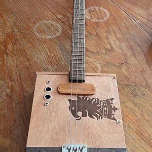May include: A cigar box guitar with a light wood body, a long neck, and three metal strings. The box features a dark brown lion logo, a wooden bridge, four silver knobs, and a blue label with the text La Gran Fabrica Drew Estate and Undercrown.