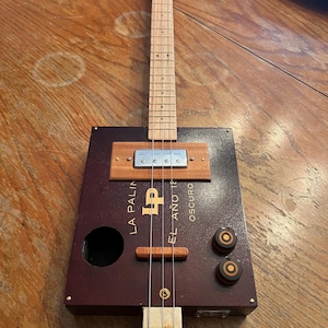 Puede incluir: Una guitarra artesanal de caja de cigarros con un cuerpo burdeos oscuro y un mástil de madera. La guitarra tiene una pastilla, clavijas de afinación y cuerdas. El texto en el cuerpo dice "LA PALIN" y "EL AÑO 18 OSCURO".