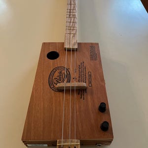 Puede incluir: Guitarra hecha a mano con una caja de cigarros de madera con el logo "Padron". Tiene un mástil de madera, tres cuerdas y dos perillas de afinación negras. La longitud total de la guitarra es de aproximadamente 76 cm.