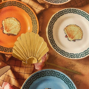 Puede incluir: Cuatro platos decorativos con un diseño de borde de clave griega. Cada plato presenta una concha estilizada en el centro. Los platos son en tonos de naranja, rosa, blanco y azul. Una concha de metal dorado está en primer plano.