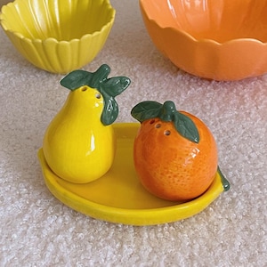 Puede incluir: Un juego de salero y pimentero de cerámica amarilla en forma de pera y naranja sobre una bandeja amarilla con forma de plátano. Los shakers tienen hojas verdes y están sobre una superficie blanca y peluda.