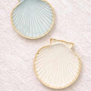 Pode incluir: Dois pratos de cerâmica em forma de concha. Um é azul claro, o outro branco. Ambos têm uma borda dourada. Os pratos têm uma textura canelada, imitando a forma natural de uma concha. São colocados sobre uma superfície branca estampada.