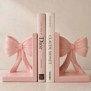 May include: A pair of light pink L-shaped bookends, each featuring a sculpted bow design on the outer side. Two books are positioned between the bookends: a pink book titled Little Book of Dior by Karen Homer and a white book titled Claude Monet by Christoph Heinrich.