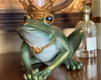 May include: A green frog figurine candle holder featuring a gold-painted crown on its head and a gold beaded necklace around its neck. The frog is in a seated position with a light pink taper candle inserted into the top of the crown.