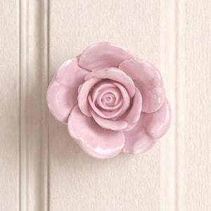 Peut inclure: Bouton de meuble en céramique rose clair en forme de rose. Le bouton est monté sur un panneau crème avec des rainures verticales. La rose a des pétales détaillés et une finition brillante, ajoutant une touche d'élégance florale aux meubles.