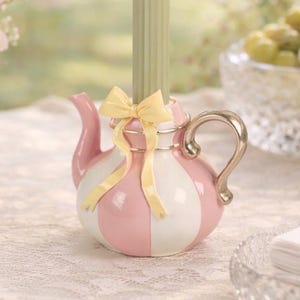 Può includere: Un portacandele decorativo a forma di teiera, con strisce rosa e bianche, un manico dorato e un fiocco giallo. Una candela verde è inserita nella parte superiore. Il portacandele è su una tovaglia di pizzo bianco.