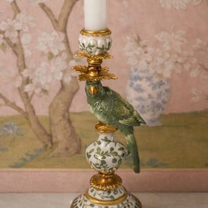 Op de afbeelding: Een decoratieve kandelaar met een groene papegaai op een wit en gouden voetstuk. De voet heeft een bloemenontwerp met gouden accenten. Bovenop de houder staat een witte kaars.