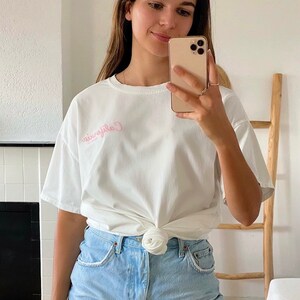 K&ouml;nnte beinhalten: Ein wei&szlig;es T-Shirt mit einem rosa gestickten Design, auf dem vorne "California" steht. Das Shirt ist in der Taille gebunden und zu einer hellblauen Jeans-Shorts kombiniert.