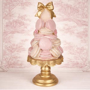 May include: A decorative macaron tower in shades of pink and white, topped with a gold bow. The tower sits on a gold pedestal, with a pink and white floral background. The macarons are arranged in tiers, creating a visually appealing dessert display.