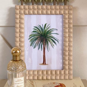 Puede incluir: Un marco de fotos beige con una impresión de palmera, coronado con pequeñas esculturas de palmeras. Un frasco de perfume con tapa dorada y el texto "Aqua Allegoria Nettare di Sole Guerlain" está delante. También se ve un libro con la palabra "Patisserie".