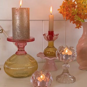 May include: A collection of lit candles in various glass candle holders. The holders are in shades of pink, yellow, and clear glass. A large, gold-glittered pillar candle sits atop a pink holder. Orange flowers in a pink vase are in the background.