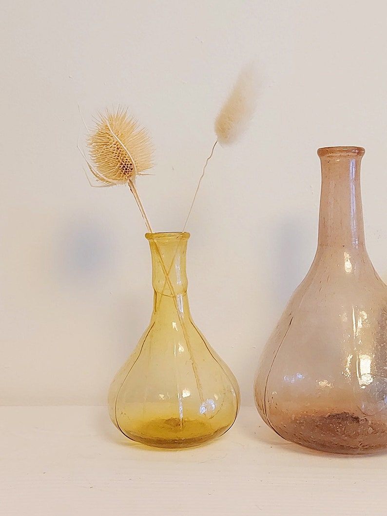 May include: Two glass vases, one amber and one clear, with dried flowers. The amber vase is smaller and has a rounder shape. The clear vase is taller and has a more elongated shape.