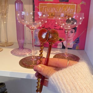 May include: Two amber-colored, glass corkscrews with spiral designs are held in front of two champagne glasses. The glasses are pink with pearl accents. A pink book with the text "TASCHEN'S NEW YORK" is in the background.