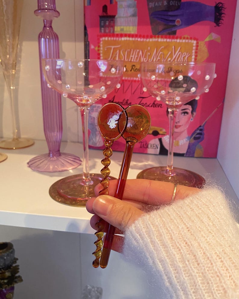May include: Two amber-colored, glass corkscrews with spiral designs are held in front of two champagne glasses. The glasses are pink with pearl accents. A pink book with the text "TASCHEN'S NEW YORK" is in the background.