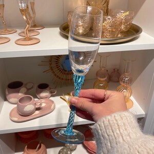 May include: A clear champagne flute with a twisted, light blue stem and base. The glass is held in front of a shelf displaying other glassware and ceramic pieces in shades of pink and peach.