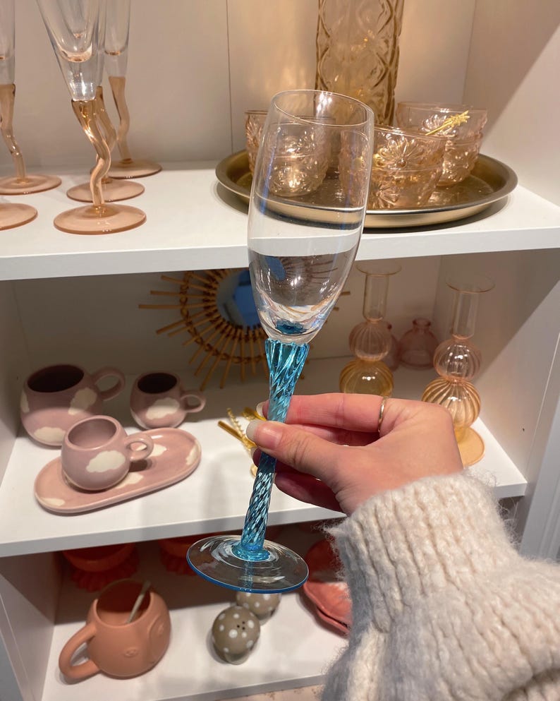 May include: A clear champagne flute with a twisted, light blue stem and base. The glass is held in front of a shelf displaying other glassware and ceramic pieces in shades of pink and peach.