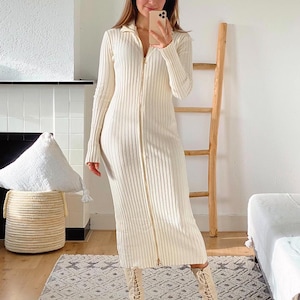 Puede incluir: Un vestido largo de punto de canalé blanco con cremallera en la parte delantera. El vestido es ajustado y tiene cuello. La modelo lleva el vestido con botas blancas de cordones.