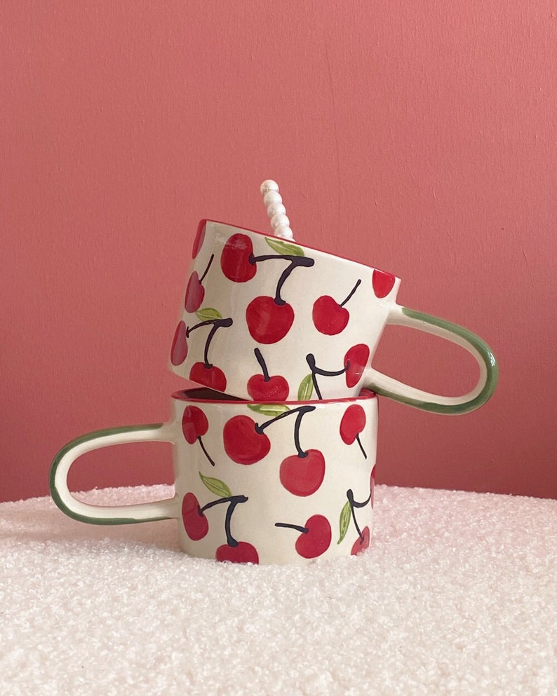 May include: Two ceramic mugs stacked on top of each other. Both mugs have a white background with a red cherry pattern. The top mug has a green handle and the bottom mug has a green rim.