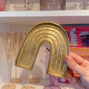 May include: A gold-colored, arched decorative tray in the shape of a rainbow. The tray has a textured surface with horizontal lines. The background includes glassware and a shelf.