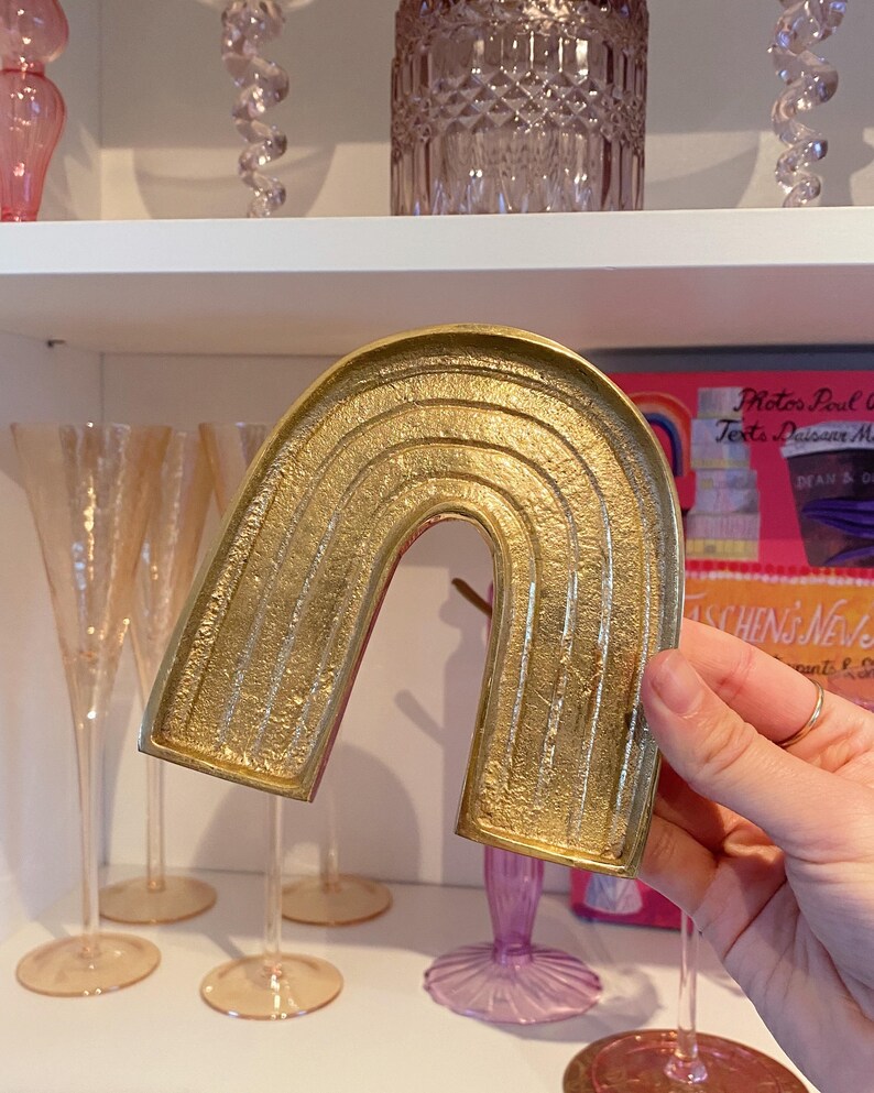 May include: A gold-colored, arched decorative tray in the shape of a rainbow. The tray has a textured surface with horizontal lines. The background includes glassware and a shelf.