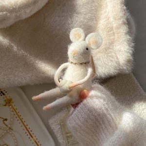 May include: A handcrafted white felt mouse figurine with a small teapot. The mouse has large ears, a pink nose, and a gold-colored necklace. The mouse is holding a small white teapot. The background is a soft, white, fuzzy material.