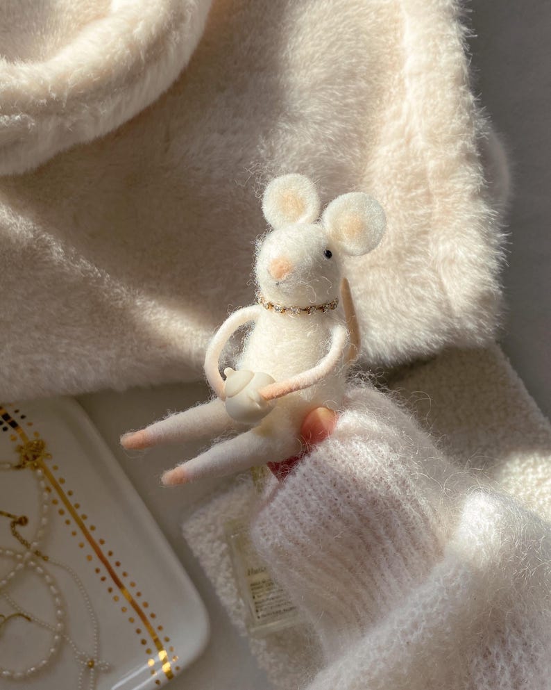 May include: A handcrafted white felt mouse figurine with a small teapot. The mouse has large ears, a pink nose, and a gold-colored necklace. The mouse is holding a small white teapot. The background is a soft, white, fuzzy material.