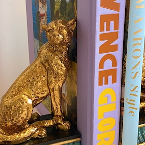 K&ouml;nnte beinhalten: Ein goldfarbener Geparden-Buchst&uuml;tze steht neben B&uuml;chern. Die Buchst&uuml;tze ist mit Flecken und einer strukturierten Oberfl&auml;che versehen. Die B&uuml;cher haben Titel in orangefarbenen und goldenen Buchstaben. Der Sockel der Buchst&uuml;tze ist t&uuml;rkis und schwarz.