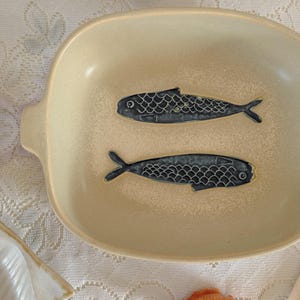 May include: Cream-colored ceramic serving dish with two dark blue fish designs. The oval dish has small handles on each side. A seashell is visible in the bottom right corner. The dish is on a white lace tablecloth.
